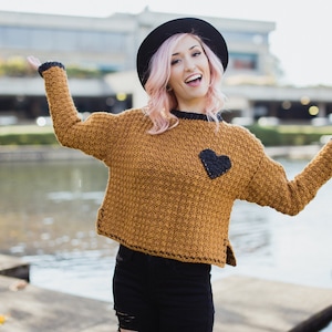 May include: A woman wearing a brown crocheted sweater with a black heart applique, a black hat, and black pants. She is smiling and has pink hair.