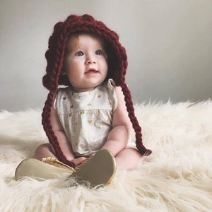May include: A baby wearing a burgundy knitted hat with long braids and gold shoes sits on a white fluffy rug.