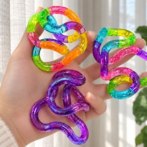 May include: Three translucent, interlocking fidget toys made of connected curved segments. One toy features a rainbow gradient of pink, orange, yellow, green, and blue. The second is a mix of blue, green, and purple. The third is solid purple.