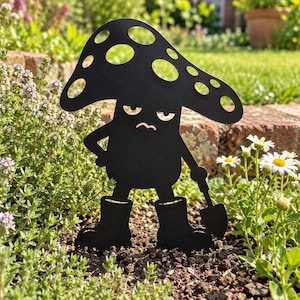 Puede incluir: Un adorno de jardín de metal negro con forma de hongo antropomórfico, con un sombrero ancho y moteado, expresión facial gruñona, brazos y piernas. La figura lleva botas y sostiene una pequeña pala en una de sus manos.