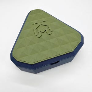 Peut inclure: Une boîte de rangement triangulaire avec une base bleu foncé et un couvercle vert olive mat. Le couvercle présente un motif pyramidal géométrique et un logo central en relief composé d'une lettre M stylisée intégrée à une forme géométrique.