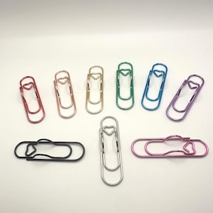 May include: A collection of eight metal paper clips in various colors, including red, rose gold, gold, green, blue, purple, black, and silver. Each paper clip features a small heart-shaped wire detail at the top, designed for holding documents.