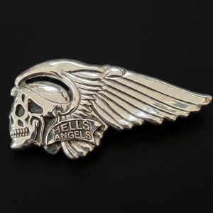 Peut inclure: Une broche en métal argenté poli représentant un crâne stylisé portant un casque ailé. Une bannière sur la partie inférieure du motif affiche le texte HELLS ANGELS en lettres capitales en relief. La pièce présente une finition métallique.