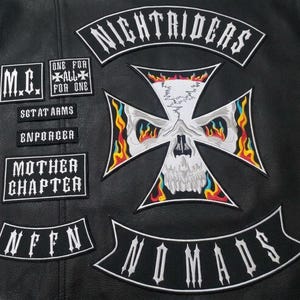 Puede incluir: Una chaqueta de cuero negro con parches bordados que incluyen una gran cruz con forma de calavera con llamas coloridas, un parche curvo Nightriders y parches rectangulares con el texto M.C., One For All For One, Sgt At Arms, Enforcer, Mother Chapter, NFFN y Nomads.