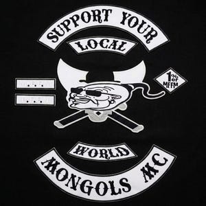 Pode incluir: Um fundo de tecido preto com emblemas bordados. As faixas superior e inferior dizem SUPPORT YOUR LOCAL e MONGOLS MC WORLD. Um emblema central mostra uma figura de desenho animado com óculos de sol e chapéu sobre machados cruzados.