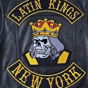 May include: A black leather vest featuring embroidered patches. An arched top patch reads LATIN KINGS in yellow. A central patch depicts a stylized skull wearing a yellow and red crown. An arched bottom patch reads NEW YORK in yellow.