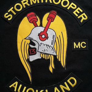 May include: A black fabric patch featuring an embroidered design of a silver helmet with a red number 13, yellow wings, and two red pistons. The text STORMTROOPER is arched above, MC is on the right, and AUCKLAND is arched below in yellow letters.