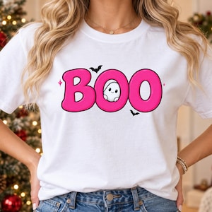 Puede incluir: Una camiseta blanca con la palabra BOO en letras grandes de color rosa brillante. Un pequeño y lindo fantasma blanco con una cara sonriente se asoma desde el centro de la letra O. Dos pequeños murciélagos negros y dos estrellas rosas rodean el texto.