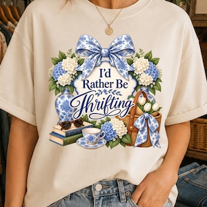 Op de afbeelding: Een crèmekleurig t-shirt met een grafische print van blauw-witte bloemenvazen, een rieten mand met witte tulpen, een stapel van twee boeken met een schildpad-zonnebril en een theekopje. De tekst luidt I'd Rather Be Thrifting.