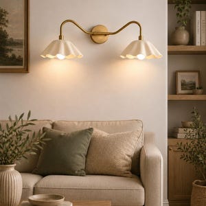 May include: A double wall sconce featuring a brass-toned metal fixture with two curved arms and two off-white, scalloped, bell-shaped shades. The light fixture is mounted on a wall and emits a warm glow from the two light bulbs inside the shades.