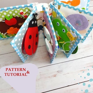 Peut inclure: Un livre en tissu avec plusieurs pages ornées d'applications en feutrine, dont une coccinelle rouge, une grenouille verte, un escargot violet et une pomme. Les pages sont bordées d'un tissu bleu clair à pois blancs. Texte : PATTERN & TUTORIAL.