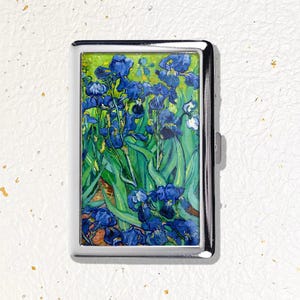 May include: A rectangular metal cigarette case featuring a reproduction of Vincent van Gogh's Irises painting. The case has a polished silver-tone frame and a hinged lid, displaying vibrant blue iris flowers with green stems and leaves.