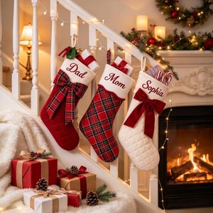 Può includere: Tre calze di Natale appese a una ringhiera bianca. La prima è rossa con un fiocco scozzese e la scritta 'Dad'. La seconda è a quadri rossi con la scritta 'Mom'. La terza è color crema con un fiocco rosso e la scritta 'Sophia'.
