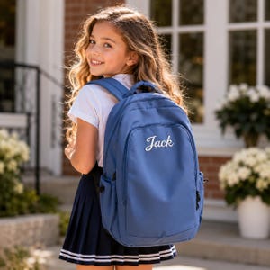 Könnte beinhalten: Ein blauer Rucksack mit dem Namen Jack in weißer Schreibschrift auf der Vordertasche. Der Rucksack verfügt über verstellbare Schultergurte, Seitentaschen und ein Hauptfach mit Reißverschluss, was ein funktionales Design für den Alltag bietet.