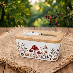 Könnte beinhalten: Eine rechteckige Butterdose aus weißer Keramik mit einer botanischen Illustration von roten Pilzen und braunem Blattwerk. Der Deckel aus hellem Holz besitzt eine Aussparung, in der ein passendes Buttermesser liegt.
