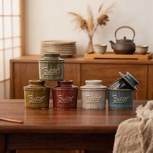 May include: A collection of five ceramic butter crocks in various colors including olive green, deep red, speckled cream, and blue. Each cylindrical container features a lid and the word Butter embossed in a cursive script on the front side.