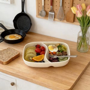 May include: A cream-colored, two-section ceramic divided plate containing sliced kiwi, cherry tomatoes, an orange slice, blueberries, a hard-boiled egg, broccoli, and sliced cucumbers, with a silver fork resting inside the dish.