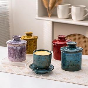 Op de afbeelding: Een verzameling van vijf keramische botervlootjes met deksels, voorzien van een glanzend, gespikkeld glazuur in de kleuren paars, mosterdgeel, dieprood en blauwgroen. Eén blauwgroen vlootje is zonder deksel getoond met bleekgele boter.
