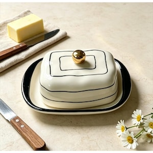 May include: A cream-colored ceramic butter dish with a rectangular base and a matching lid featuring black hand-painted line details. The lid is topped with a small, polished gold-tone spherical knob. The dish sits on a flat surface.