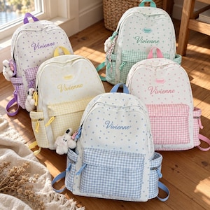 Könnte beinhalten: Fünf Rucksäcke in verschiedenen Pastellfarben, jeweils mit Sternenmuster auf dem Hauptteil, einer karierten Vordertasche und dem Namen Vivienne in Schreibschrift. Jeder Rucksack verfügt über Seitentaschen, Reißverschlüsse und einen kleinen Plüschtieranhänger.