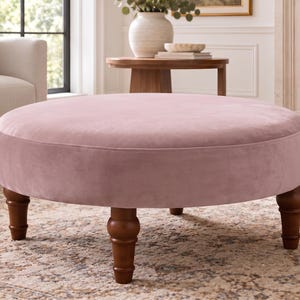 May include: A round ottoman upholstered in soft, light pink velvet fabric. The piece features a smooth, circular top surface and is supported by four dark brown wooden legs with a turned, spindle-style design that tapers toward the floor.