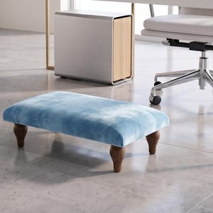May include: A rectangular footstool featuring a soft, light blue velvet-textured top cushion. The piece is supported by four short, dark brown wooden legs with a rounded, turned design, providing a stable base for resting feet while seated.