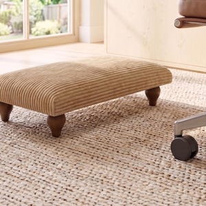 May include: A rectangular footstool upholstered in tan corduroy fabric with a ribbed texture. The footstool features four dark brown, rounded wooden feet that elevate the cushioned top section, providing a stable base for resting feet.