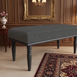 May include: A rectangular bench featuring a dark grey upholstered seat with a textured, speckled pattern. The bench is supported by four turned, matte black wooden legs that feature decorative ring details near the top of each leg.