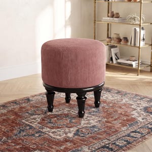 Puede incluir: Un taburete redondo tapizado en tela de pana de color rosa empolvado con una textura de rayas verticales. El asiento se apoya sobre cuatro patas de madera torneada de color marrón oscuro con un acabado brillante y un diseño clásico.