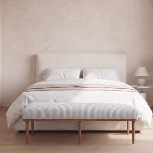 May include: A bedroom scene featuring a white upholstered bed with a matching headboard, white bedding, and a beige throw blanket. A light blue and white striped bench with wooden legs sits at the foot of the bed. A small lamp sits on a side table.