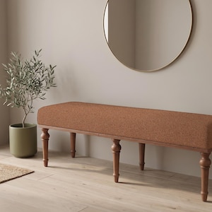 May include: A long, rectangular bench featuring a textured, rust-colored upholstered seat cushion supported by a wooden frame with six turned legs. The bench has a simple, elongated design with a consistent fabric finish across the top surface.
