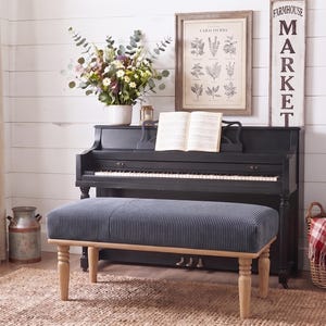 May include: A rectangular bench with a dark blue corduroy upholstered seat and four light-colored wooden legs. The bench is positioned in front of a black upright piano with an open music book resting on the music stand and a floral arrangement.