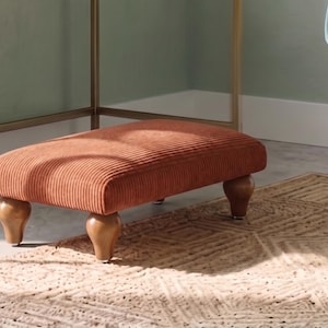 Puede incluir: Un reposapiés rectangular tapizado en tela de pana de color óxido con una textura acanalada distintiva. La pieza cuenta con cuatro patas de madera redondeadas y bulbosas con un acabado liso y pulido, que proporcionan una base estable.