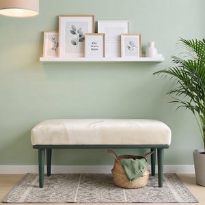 May include: A cream-colored upholstered bench with dark green legs sits on a patterned rug. A woven basket with a green cloth, a potted palm plant, a white wall shelf with five framed prints, and a hanging canvas tote bag are present.