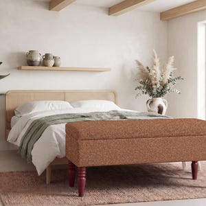 Puede incluir: Un banco de almacenamiento rectangular tapizado en una tela texturizada de color naranja quemado. El banco cuenta con cuatro patas de madera torneada de color rojo oscuro y una tapa con bisagras, situado a los pies de una cama con cabecero de rejilla.