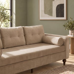 May include: A beige velvet sofa featuring two large tufted back cushions, a single long seat cushion, and a cylindrical bolster pillow on the right side. The sofa is supported by dark brown wooden legs and has a smooth, uniform fabric texture.