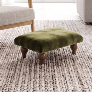 Puede incluir: Un reposapiés rectangular tapizado en tela de terciopelo verde oliva. La pieza presenta una superficie superior capitoné y se apoya sobre cuatro patas de madera de color marrón oscuro con un diseño torneado y redondeado, ofreciendo una base estable.