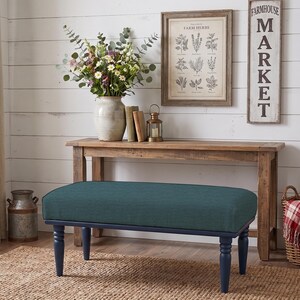 May include: A rectangular bench featuring a deep teal upholstered seat cushion supported by four dark navy blue turned wooden legs. The bench is positioned in front of a wooden console table, with a floral arrangement and books visible above.