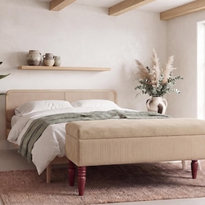 Op de afbeelding: Een rechthoekige opbergbank van beige corduroy met vier donkerrode gedraaide houten poten, geplaatst aan het voeteneinde van een lichthouten bedframe met een gevlochten hoofdbord, bedekt met wit beddengoed en een saliegroene sprei.