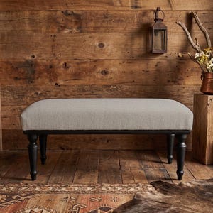 May include: A rectangular bench featuring a light grey upholstered seat cushion supported by four dark brown turned wooden legs. The bench has a clean, simple design with a smooth fabric top and sturdy, rounded legs that provide a stable base.