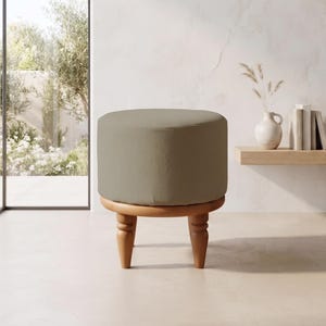 Puede incluir: Un taburete redondo con un cojín de asiento tapizado en color verde oliva, sostenido por cuatro patas de madera torneada con un acabado marrón cálido. El taburete tiene forma cilíndrica y se asienta sobre un suelo liso y claro.