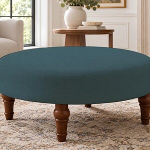 May include: A round ottoman upholstered in a solid teal fabric. The piece features a low-profile circular cushion design supported by four turned wooden legs with a medium brown finish, providing a stable base for the furniture item.