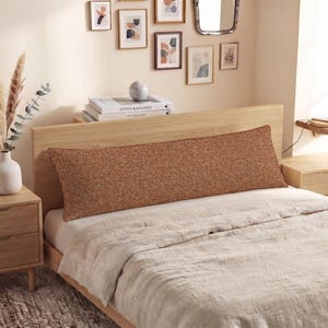 May include: A long, rectangular body pillow featuring a textured, burnt orange fabric cover. The pillow is positioned horizontally across the head of a bed, resting against a light-toned wooden headboard, providing a solid color accent to the bedding.