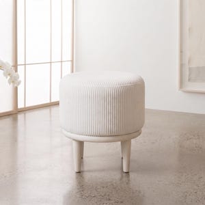 May include: A round, off-white ottoman featuring a vertically ribbed, corduroy-like fabric texture on the seat. The piece is supported by four short, tapered, light-colored wooden legs connected by a circular frame base beneath the cushion.