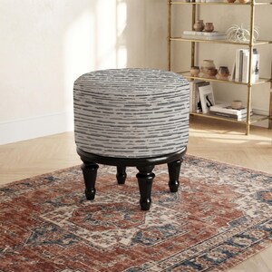 May include: A round ottoman featuring a textured white and gray horizontal striped fabric upholstery. The piece is supported by four dark brown, turned wooden legs that provide a stable base for the circular, cushioned seat top.