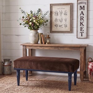 May include: A rectangular bench with a dark brown velvet upholstered seat and four turned navy blue wooden legs. The bench is positioned in front of a wooden console table, which holds a ceramic vase with flowers, two books, and a small lantern.