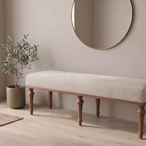 Puede incluir: Un banco rectangular largo con un cojín de asiento tapizado en color crema con rayas verticales, sostenido por seis patas de madera torneada en un acabado marrón medio. El banco tiene una parte superior acolchada y una estructura de madera robusta.