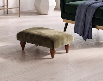 May include: A rectangular footstool upholstered in olive green velvet fabric, featuring four rounded, dark brown wooden legs. The footstool has a low-profile design with a soft, plush surface texture and is positioned on a light-colored wood floor.