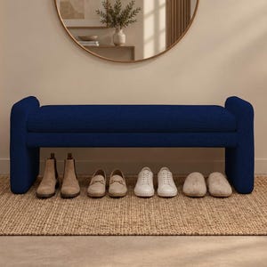 May include: A navy blue upholstered bench with rounded ends and a flat seat. The bench is positioned over a rectangular woven jute rug. Four pairs of shoes, including tan boots, beige loafers, white sneakers, and beige slippers, are placed underneath.