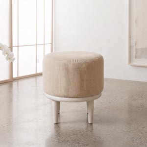 May include: A round ottoman featuring a beige corduroy upholstered seat with a vertical ribbed texture. The seat is supported by a circular frame and four short, tapered legs finished in a smooth, light-coloured wood or painted material.